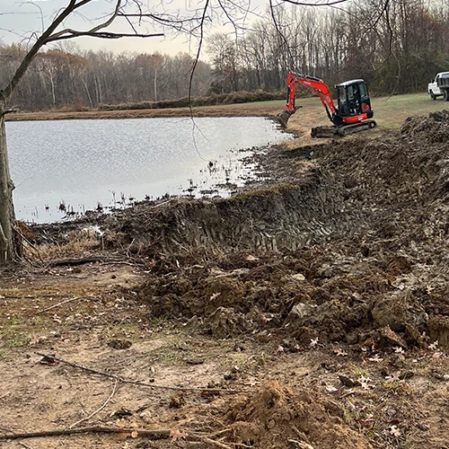 A backhoe Excavator is trenching next to a pond to enlarge it